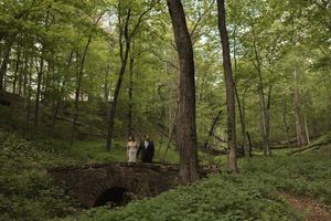 Bride and groom stand romantically on stone arch bridge surrounded by lush green forest at wedding.