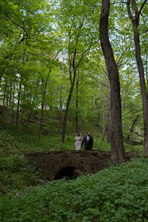 Bride and groom stand on rustic stone arch bridge surrounded by vibrant green woodland trees.