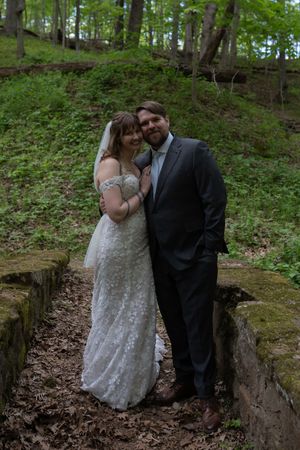 Bride and groom posing together against mossy stone wall in lush green forest.
