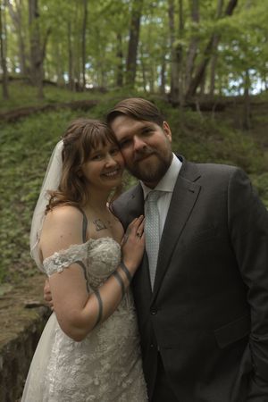 Smiling bride and bearded groom pose together in lush green forest wedding portrait.