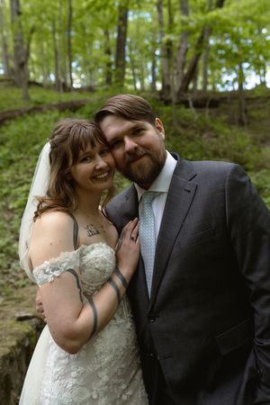 Smiling bride and groom pose together in a lush green forest, with bride in lace gown and groom in gray suit.