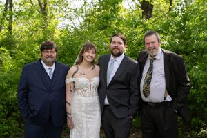 Bride in lace gown poses with groom and two men in suits outdoors at wedding.