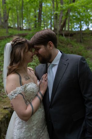 Bride and groom touch foreheads tenderly in romantic woodland portrait, both dressed in wedding attire.