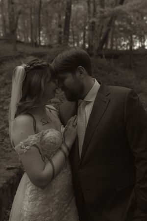 Black and white photo of a bride and groom touching foreheads in a romantic forest wedding portrait.