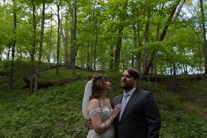 Newlywed couple poses romantically in a lush green forest setting, bride in lace gown with veil.