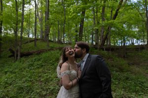 Groom kisses laughing bride on cheek during joyful outdoor wedding portrait in lush green woods.
