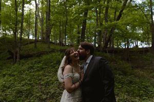 Groom kisses laughing bride on cheek in romantic outdoor forest wedding portrait.