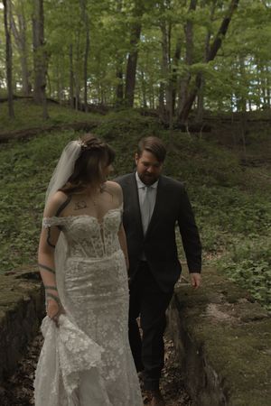 Bride in lace off-shoulder wedding gown and groom in dark suit walking through a lush green forest.