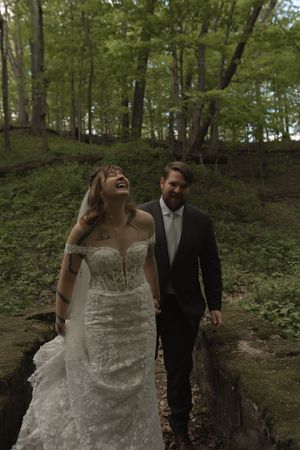 Laughing bride in lace gown and groom in dark suit walk together through scenic woodland setting.