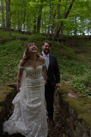 Laughing bride in lace gown holding grooms hand walking along stone path through green woodland setting.