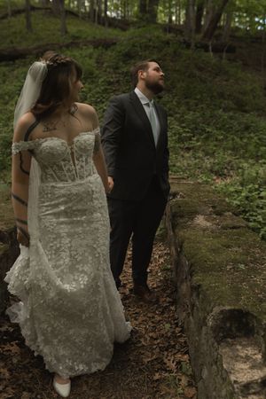 Bride in floral lace gown and veiled groom in dark suit exploring a scenic wooded stone path.