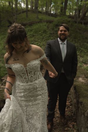 Bride in lace off-shoulder gown holds groom's hand walking through lush green forest during outdoor wedding.