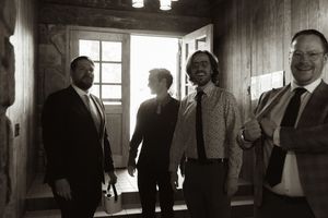 Black and white photo of four men in suits smiling together in a warmly backlit wooden doorway.