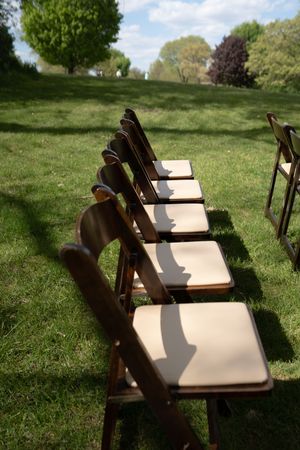 Row of wooden folding chairs with cream cushions set up on green grass for outdoor wedding ceremony.