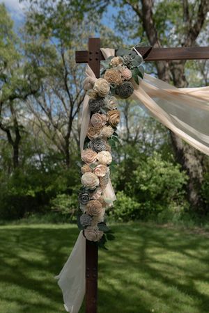 Wooden wedding cross decorated with cream and sage paper flowers and flowing ivory ribbon in an outdoor garden setting.