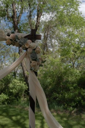 Rustic wooden cross wedding arch adorned with cream and sage roses with flowing ribbon in garden.