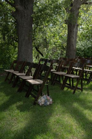 Rows of dark wooden folding chairs set up for an outdoor wedding ceremony under large shady trees.