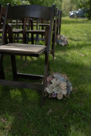 Pastel floral bouquet resting on green grass beside dark wooden folding chairs at outdoor wedding.