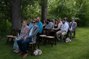 Wedding guests seated on wooden chairs outdoors under shaded trees during garden ceremony.