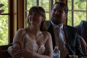 Bride with tattoos in lace dress and groom in suit smiling during wedding reception at wooden table by window.