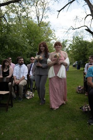 Bridesmaid in pink gown escorted down outdoor wedding aisle by woman in gray suit holding bouquet.