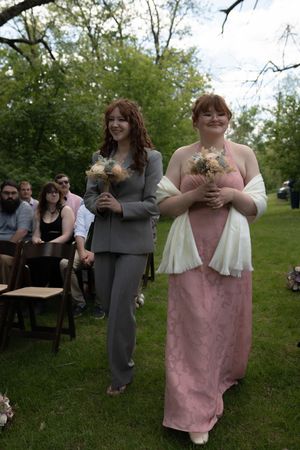 Two bridesmaids walking down outdoor aisle carrying bouquets in gray pantsuit and pink gown with white shawl.