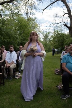 Bridesmaid in lavender gown walks down outdoor wedding aisle holding bouquet before seated guests.
