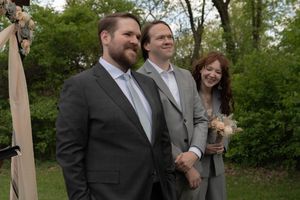 Two groomsmen in gray suits smile with red-haired bridesmaid in gray suit at outdoor wedding ceremony.