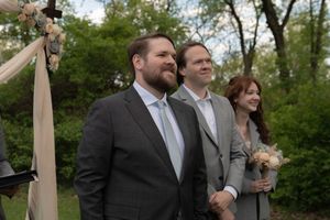 Groom in gray suit smiles at outdoor woodland wedding ceremony beside officiant and bridesmaid.