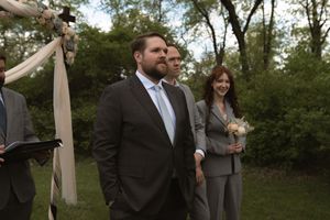 Groom in dark suit smiles at outdoor wedding ceremony with officiant and bridesmaid in background.