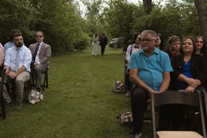 Wedding guests seated in wooden chairs watch an outdoor garden ceremony among trees.