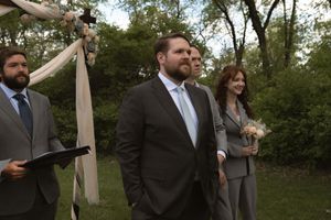 Groom in dark suit standing at outdoor wedding altar with wedding party beside floral arch.