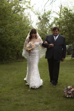 Father walking bride in lace mermaid wedding dress down outdoor grass aisle holding bouquet.