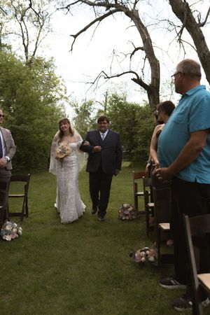 Bride walks down outdoor aisle with escort past seated guests during garden wedding ceremony.