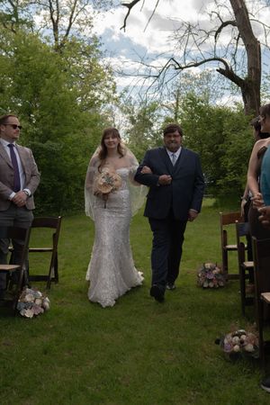 Smiling bride in lace gown walks down grass aisle with escort during outdoor wedding ceremony.
