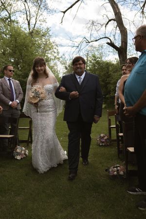 Bride in white lace gown and veil walks down outdoor aisle with escort past seated wedding guests.