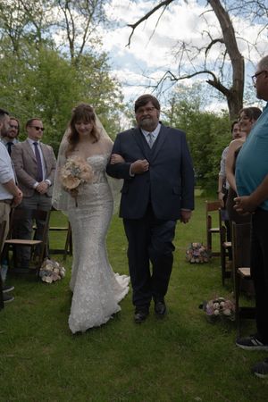 Bride in lace gown walks down outdoor aisle with father during garden wedding ceremony.