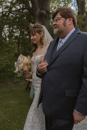 Bride in lace gown holds bouquet while walking with older gentleman in suit during wedding processional.