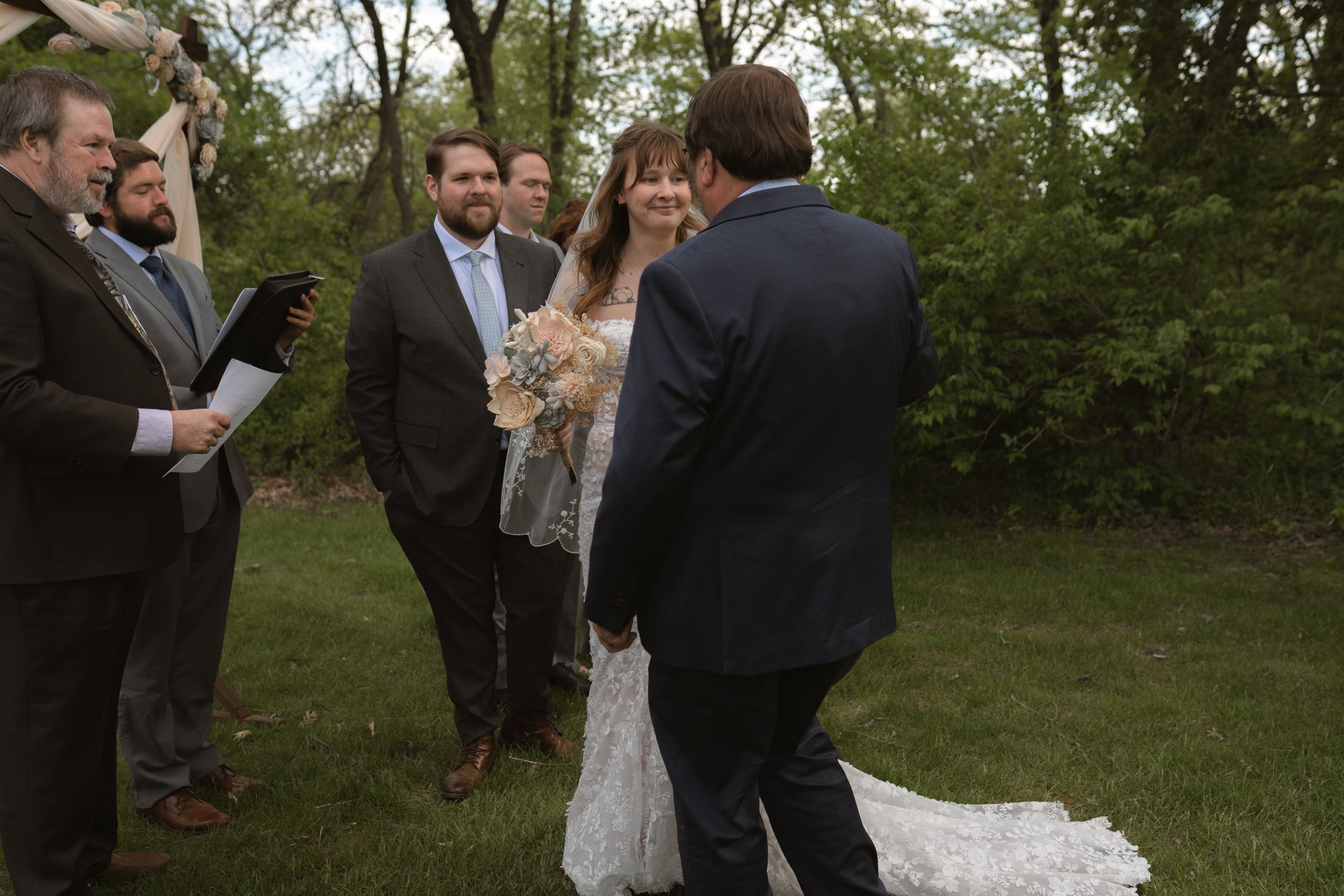 Bride smiling at groom during outdoor wedding ceremony with guests and officiant standing nearby.