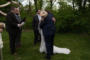 Outdoor wedding ceremony with bride and groom embracing, officiant reading, lush green trees in background.