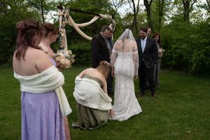 Bridesmaids assist bride in white lace gown and veil during outdoor garden wedding ceremony under floral arch.