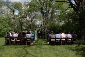 Outdoor wedding ceremony with guests seated on wooden chairs facing couple at floral arch.
