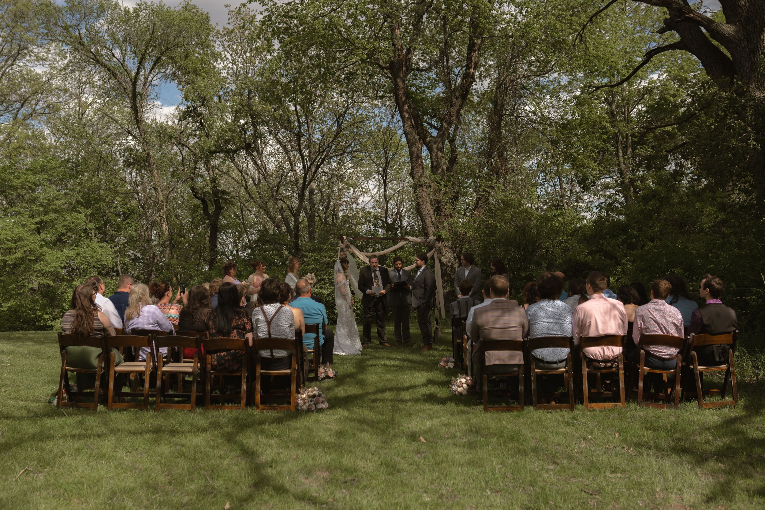 Outdoor wedding ceremony with guests seated on wooden chairs in a lush green garden surrounded by trees.