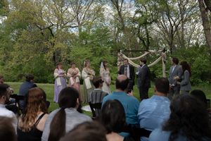 Wedding guests seated outdoors watch couple exchange vows under rustic floral arch surrounded by lush green trees.