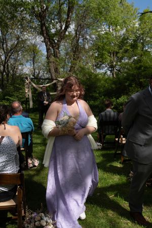 A bridesmaid in a lavender halter gown with white wrap carries a bouquet while walking down the outdoor wedding aisle.