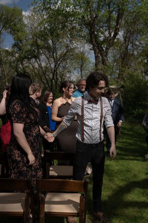 Groom in suspenders and bow tie walks down outdoor wedding aisle holding hands with guest amid lush green trees.