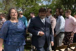 Couple in formal wedding attire smiling and walking outdoors among wedding guests on sunny day.