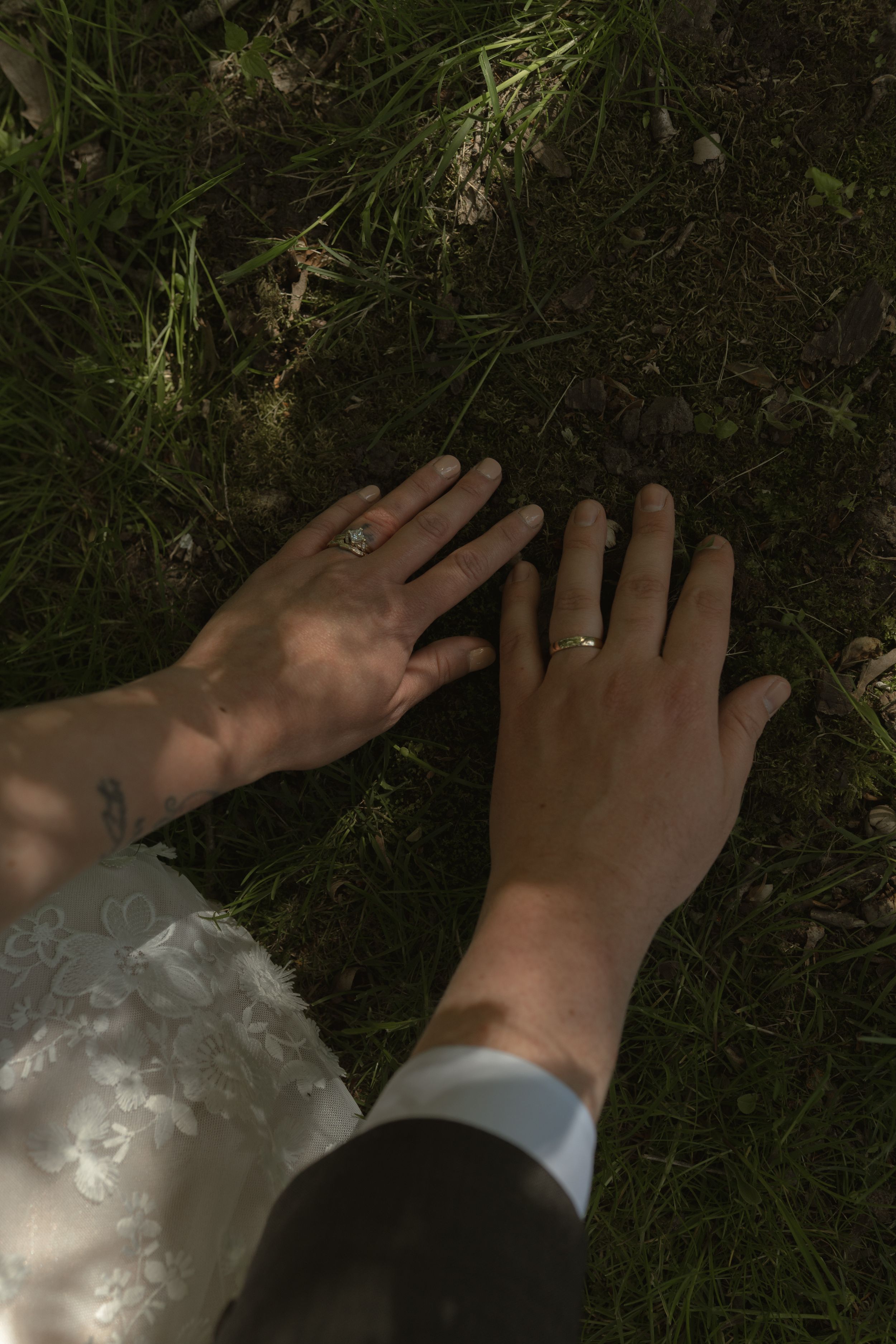 Close-up of bride and groom hands on grass showing wedding rings, bride has wrist tattoo.