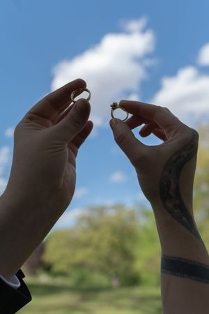 Hands holding wedding rings up against a blue sky, one wrist adorned with a tattoo.