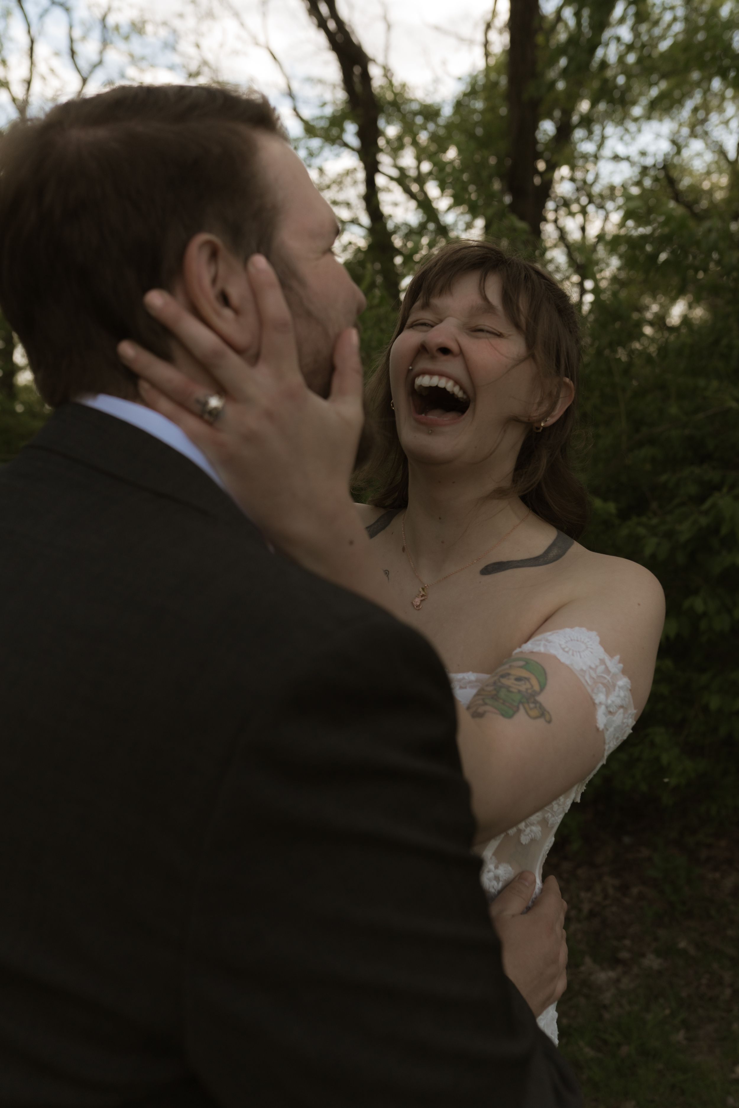 Joyful bride laughing while embracing groom outdoors, showing floral tattoo and lace wedding dress.